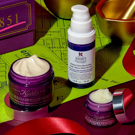 Tre Kiehl's Super Multi-Corrective produkter – en kramburk, en ogonkram och en serumflaska – ar arrangerade på ett rott band, bredvid en lila låda och guldfargade festliga dekorationer, mot en limegron bakgrund med kartmonster.