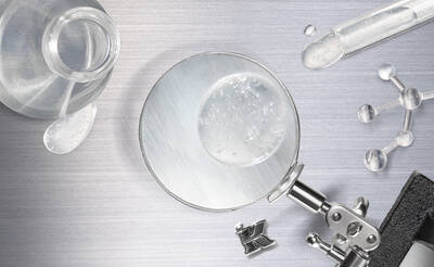 A magnifying glass focuses on a droplet of water on a metallic surface, surrounded by a glass vial, a test tube, and molecular models.