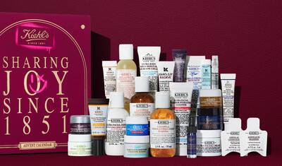Kiehl's Adventskalender presentforpackning i djuprott med guldtext, visad bredvid 24 mini hudvårdsprodukter inklusive Ultra Facial Cream, Calendula Toner, Retinol Serum och Lip Balm #1. En rosa sprayfarg-grafik på sidan framhaver setets varde.