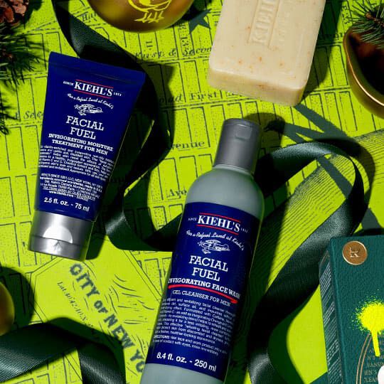 Kiehl’s Facial Fuel rengoringsmedel och fuktighetskram arrangerade med en tvål och grona juldekorationer på en ljusgul bakgrund.