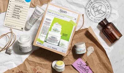 Flat lay of Kiehl's skincare products, including creams and serums, on crumpled brown paper with an ingredient list and a tag. A vintage apothecary bottle is also visible.
