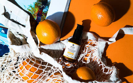 A flat lay featuring a bottle of skincare serum nestled in a cream-colored net bag, surrounded by bright oranges on an orange background, with a magazine featuring a car in the corner.
