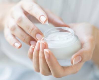 A person's hands are gently holding a small jar of cream, with one hand poised to lift the lid. The cream inside the jar is white and smooth-looking.