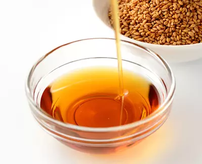 A glass bowl filled with golden brown oil, with a stream of oil being poured from above; a bowl of sesame seeds is in the background.