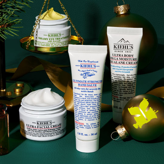 Ett festligt arrangemang av Kiehl's produkter inklusive Creamy Eye Treatment på gyllene fjall, Ultra Facial Cream, Ultimate Strength Hand Salve och Ultra Body Mega Moisture Squalane Cream, omgivet av grona ornament, granris och guldiga band på en morkgron bakgrund.
