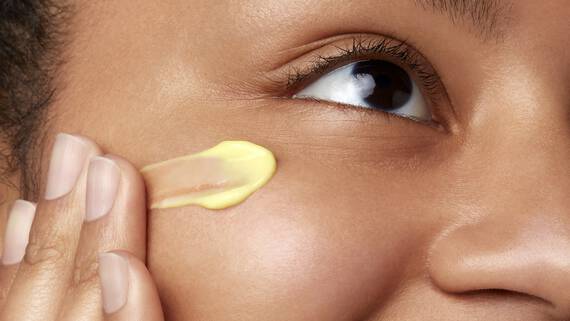 A close-up of a woman's face with a gentle smile, applying a creamy yellow moisturizer to her cheek. Her eye is focused and bright, showcasing healthy skin.