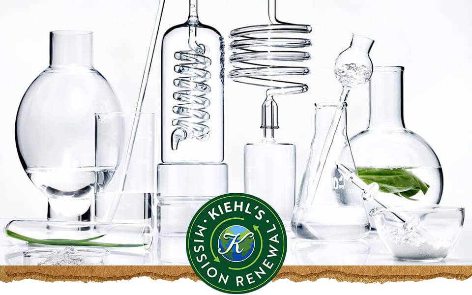 En samling laboratoriekarl i klarglas fyllda med vatten och ett gront lov med Kiehl's logotyp till hoger.