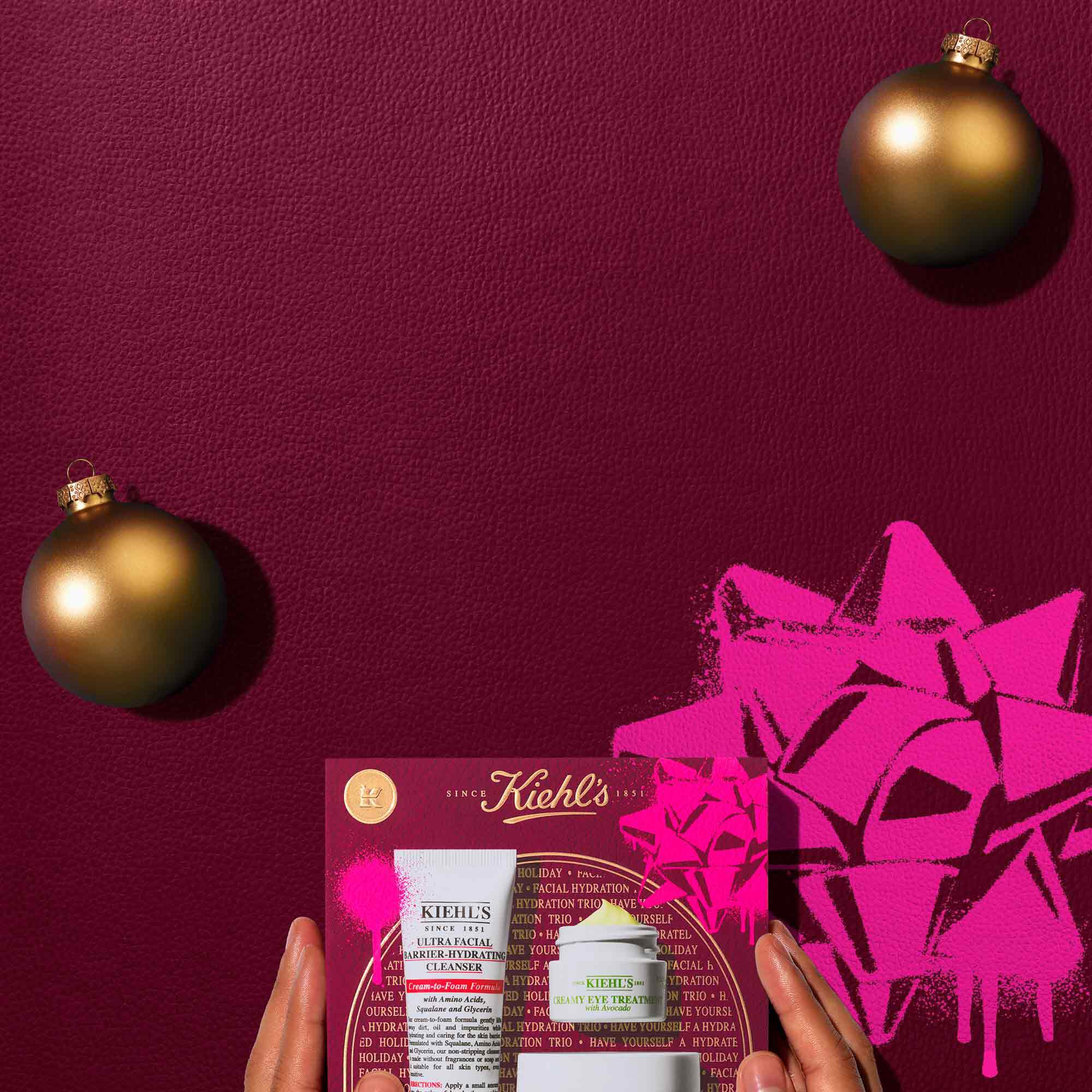 A graphic, set against a dark red textured background, features three Kiehl’s skincare products and a gift box. At the top, a white tube of "KIEHL’S ULTRA FACIAL BARRIER-HYDRATING CLEANSER", a white jar of "KIEHL’S ULTRA FACIAL CREAM", and a small open white jar of "KIEHL’S CREAMY EYE TREATMENT WITH AVOCADO" with visible pale yellow cream are displayed. Two gold spherical ornaments are also present. Below, a red Kiehl’s gift box showcasing the same three products is visible. The gift box has bright pink graphic paint splatters and a bow design.