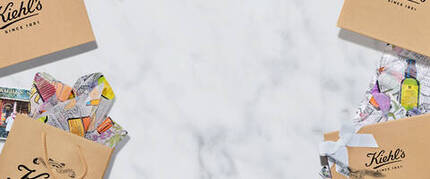 Alt-text: En flat lay-bild med Kiehl's papperspåsar och presentforpackningar på en marmorbelagd yta, omgivna av fargglada monster och en fotografi.