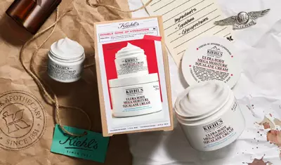 The image features Kiehl's skincare products, including various cream jars and a box labeled "Double Dose of Hydration." Surrounding items include a tag reading "Apothecary Since 1851" and a vintage-style letter with ingredients listed.