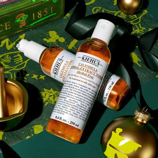 Kiehl’s Calendula Herbal-Extract Toner och serum arrangerade med guldfargade julpynt och gront band på en festlig morkgron bakgrund.