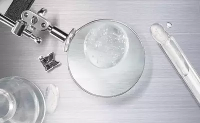 A close-up of a magnifying glass surrounded by laboratory equipment, including a test tube and a dropper, with droplets of liquid on a metallic surface.
