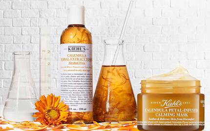 Bild av hudvårdsprodukter med olika burkar och flaskor på en vit tegelvagg. Produkterna inkluderar en ansiktskram, ett serum och en lugnande mask, alla dekorerade med ringblommor.