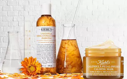 Bild av hudvårdsprodukter med olika burkar och flaskor på en vit tegelvagg. Produkterna inkluderar en ansiktskram, ett serum och en lugnande mask, alla dekorerade med ringblommor.