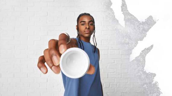 En person i blå armlos skjorta håller en liten vit skål mot kameran, som står mot en vit vagg med textur.