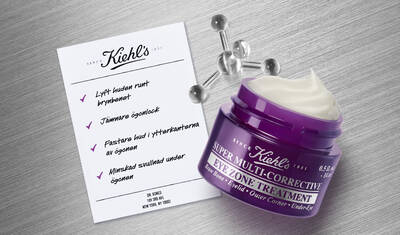 A jar of Kiehl's Super Multi-Corrective Eye Zone Treatment beside a printed list of benefits, highlighting improvements in brow area lift, eyelid smoothing, outer corner firmness, and under-eye de-puffing percentages.