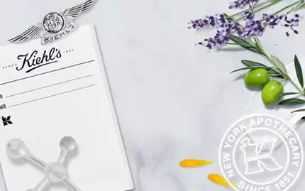 Alt-text på svenska: "Kiehl's historiska foto från deras apotek i New York, omgiven av lavendel, oliver och blomblad, med deras logotyp och text som visar grundandet 1851."
