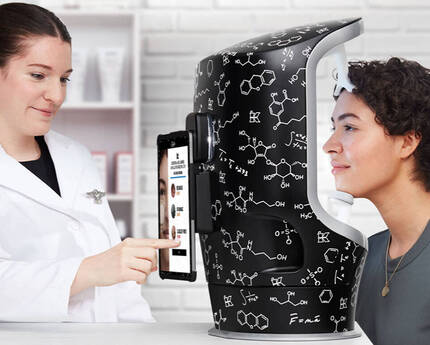 En kvinna i vit labbrock visar en digital skarm for en person som sitter vid en maskin med vetenskapliga monster.