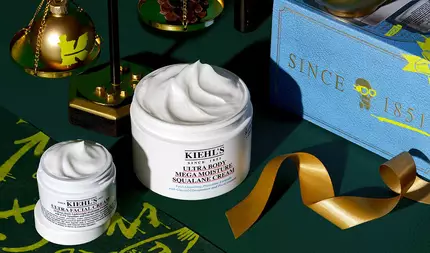 En grafik, mot en morkgron bakgrund med guldaccenter, visar två oppna vita burkar med synliga virvlar av produkt. Den mindre burken ar "KIEHL'S ULTRA FACIAL CREAM" och den storre burken ar "KIEHL'S ULTRA BODY MEGA MOISTURE SQUALANE CREAM." I bakgrunden finns en blå Kiehl's presentforpackning med guldtext som laser "SINCE 1851", ett guldband, en guldbalansvåg med en prydnad och en liten kotte.