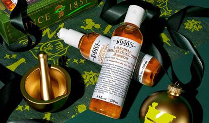 En grafik, mot en festlig morkgron bakgrund med guldaccenter, visar två flaskor: "Kiehl's Calendula Deep Cleansing Foaming Face Wash" och "Kiehl's Calendula Herbal-Extract Toner", båda med genomskinlig forpackning och barnstensfargad vatska. Scenen innehåller en mortel och mortelstot i guld, ett guldprydnadsforemål med en gron "K"-logotyp, svart band och bakgrunden dekorerad med guldillustrationer.