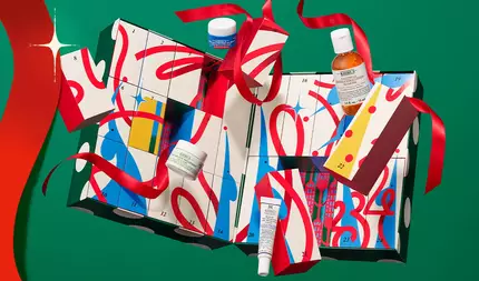 Kiehls adventskalender med numrerade presentaskar och hudvårdsprodukter, inslagen i ett rott band på en festlig gron bakgrund.