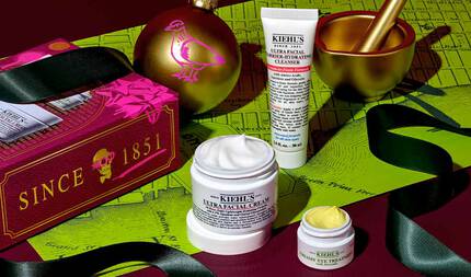 En festlig grafik som visar en vit tub med "KIEHL'S ULTRA FACIAL BARRIER-HYDRATING CLEANSER", en vit burk med "KIEHL'S ULTRA FACIAL CREAM" med synlig vit kram och en liten vit burk med "KIEHL'S CREAMY EYE TREATMENT WITH AVOCADO" med synlig ljusgul kram, allt arrangerat på en morkrod och gron bakgrund med ett kartmonster. En mortel och mortelstot i guld syns uppe till hoger. Uppe till vanster finns en julprydnad i guld med ett rosa fågelmonster och en rod presentforpackning med "SINCE 1851" och ett dodskallemonster. Svart band ar draperat runt produkterna.