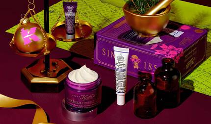 En festlig grafik mot en morkrod bakgrund med ett gront kartmonster visar flera Kiehl's-produkter och juldekorationer. En balansvåg i guld ar framtradande, med en sfarisk prydnad i guld med en rosa "K"-logotyp på ena sidan och en liten lila tub med "Kiehl's Super Multi-Corrective Eye Zone Treatment" på den andra. I forgrunden visas en oppen lila burk med "Kiehl's Super Multi-Corrective Cream" med synlig kram. En vit och lila tub med "Kiehl's Retinol Skin-Renewing Daily Micro-Dose Serum" står bredvid. I bakgrunden syns en mortel och mortelstot i guld med tallgrenar och en lila presentforpackning med "SINCE 1851" och en dodskalle. Två barnstensfargade glasflaskor och ett guldband kompletterar det festliga arrangemanget.